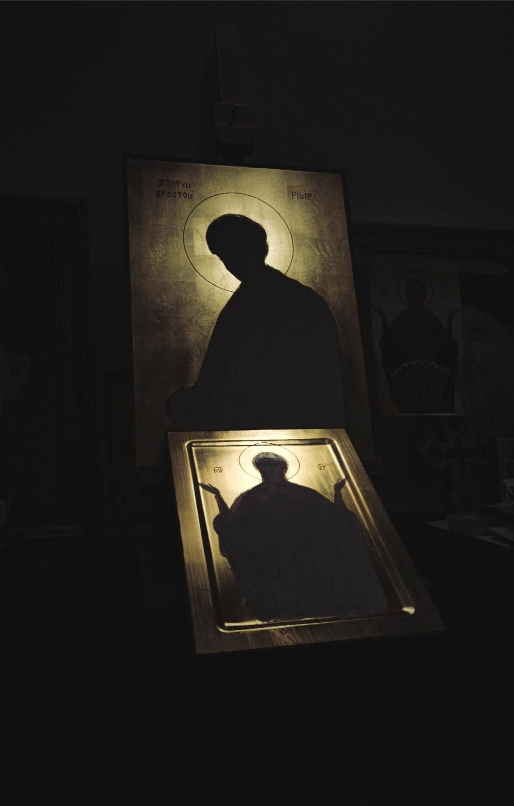 Două icoane în lucru, cu aureole și fundaluri aurii iluminate într-un spațiu întunecat.