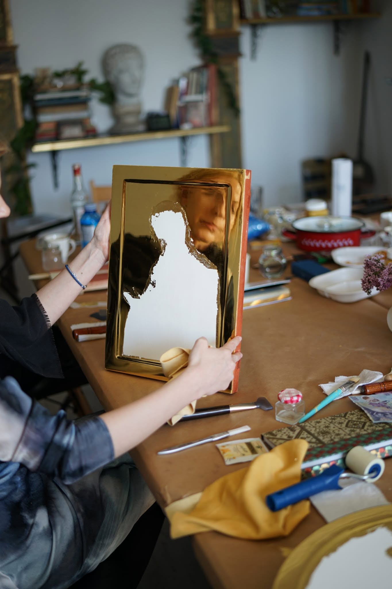 Artista ținând un panou cu fundal auriu, în fața mesei cu materiale.