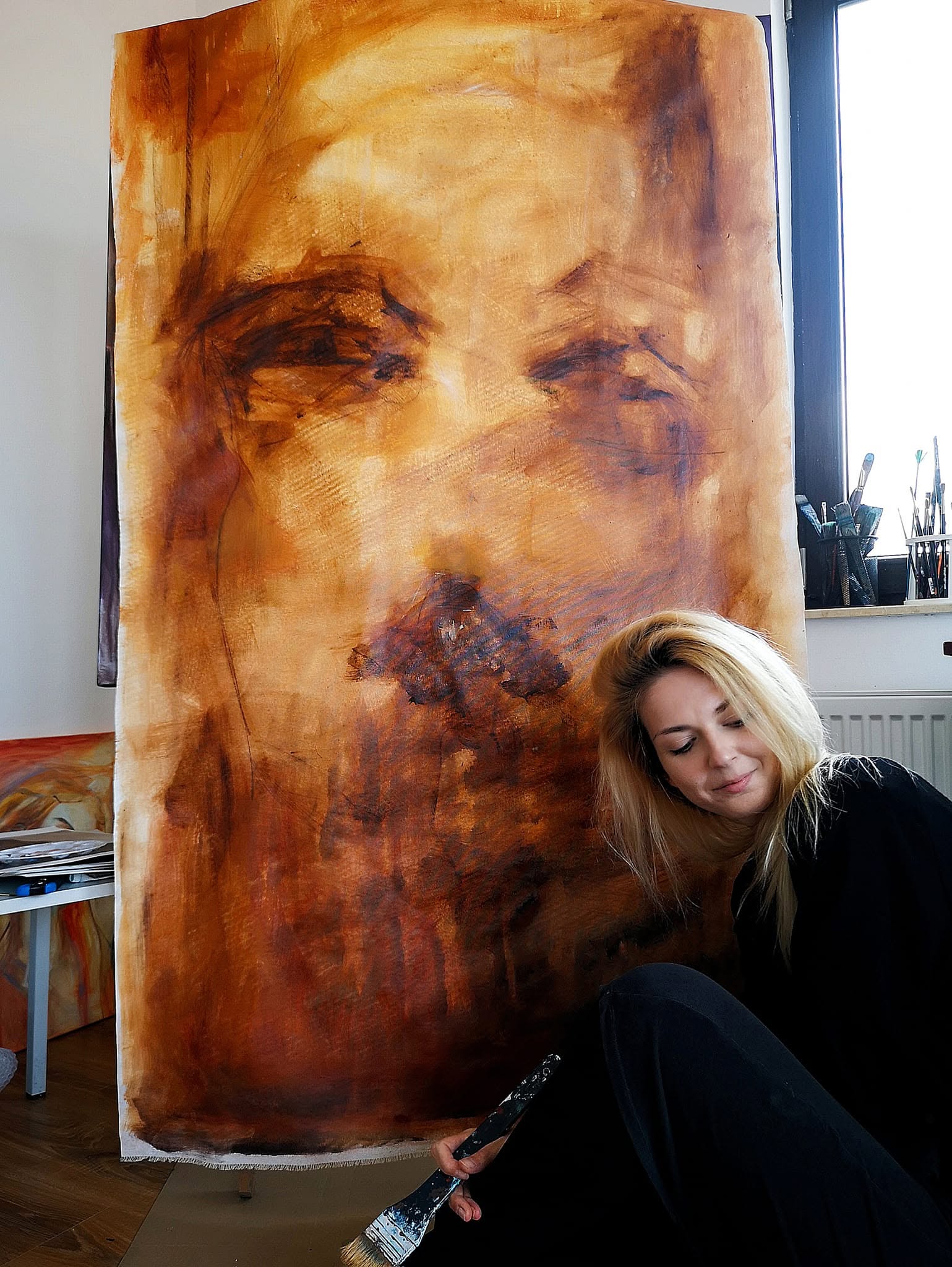 Artista cu o pensulă lată, lângă un portret de mari dimensiuni în tonuri calde.