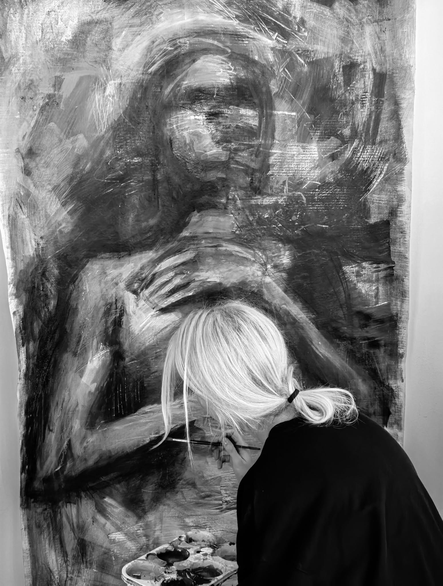 Artista, văzută din spate, lucrând în fața unei compoziții figurative.