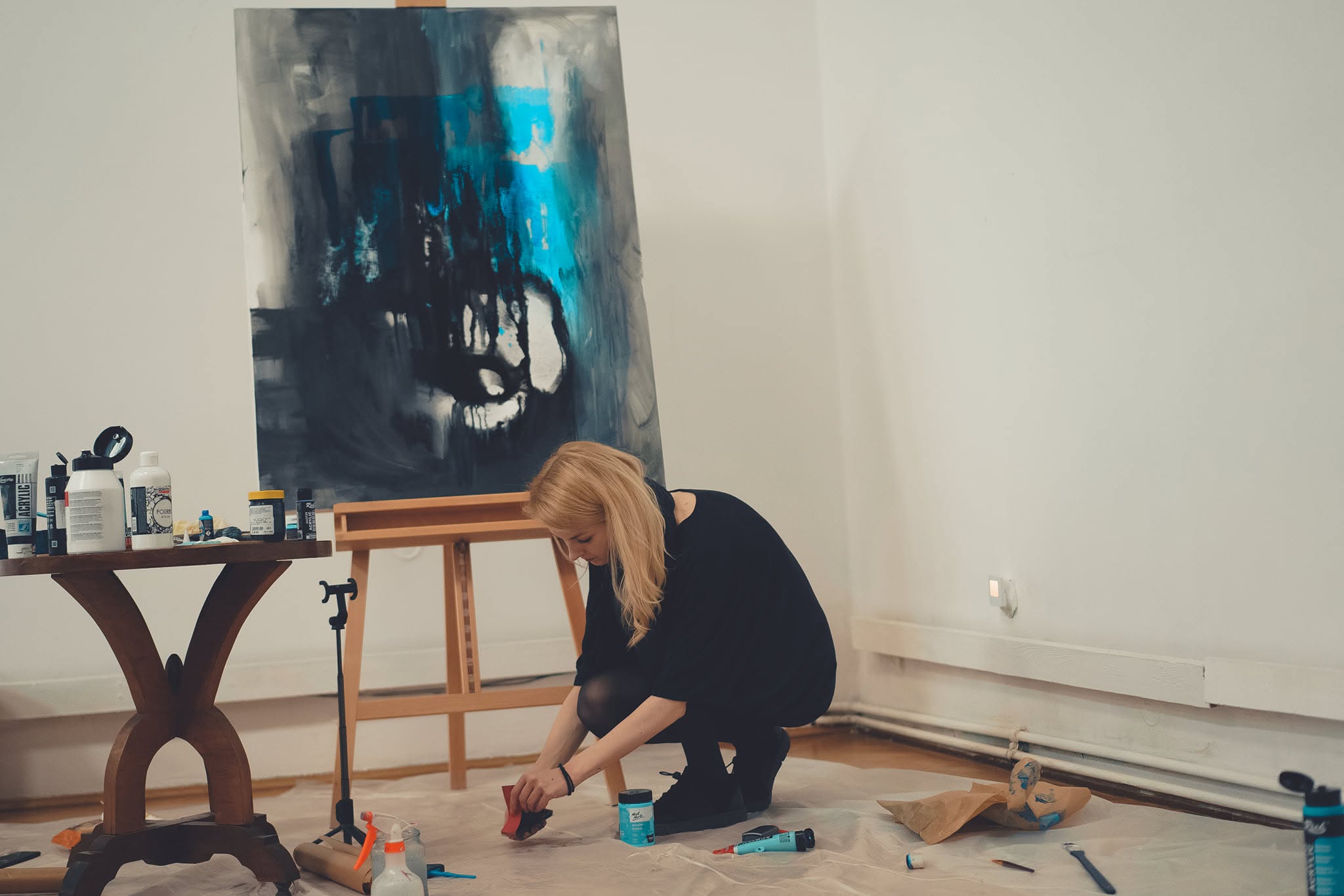 Artista pregătind culoarea în fața șevaletului, cu pânza și materialele de pictură în cadru.