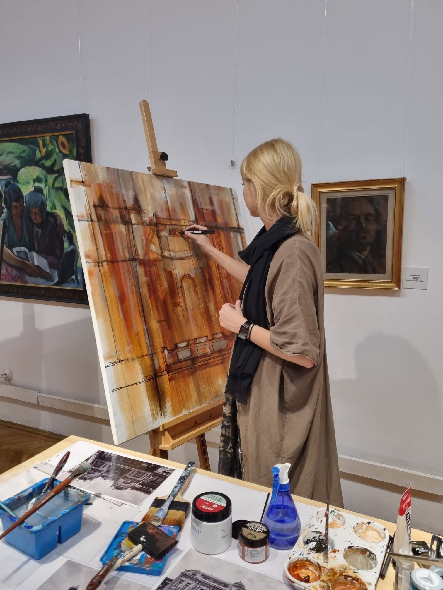 Anita Frâncu desenând pe o pânză în tonuri de ocru și brun, la șevalet, în galeriile Muzeului de Artă Brașov.